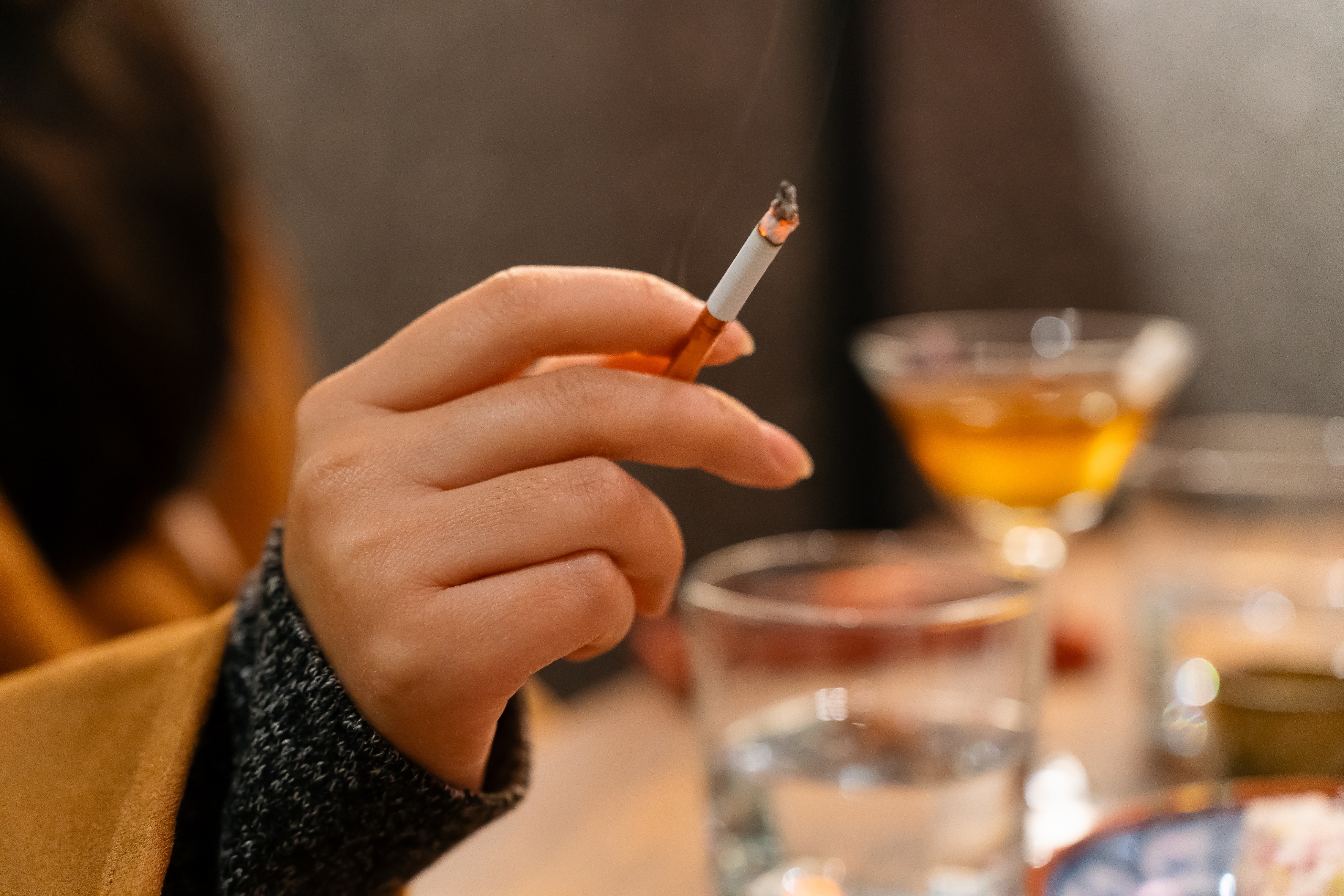 En hånd som holder en sigarett over et bord med briller, et cocktailglass og uskarpt bakgrunn