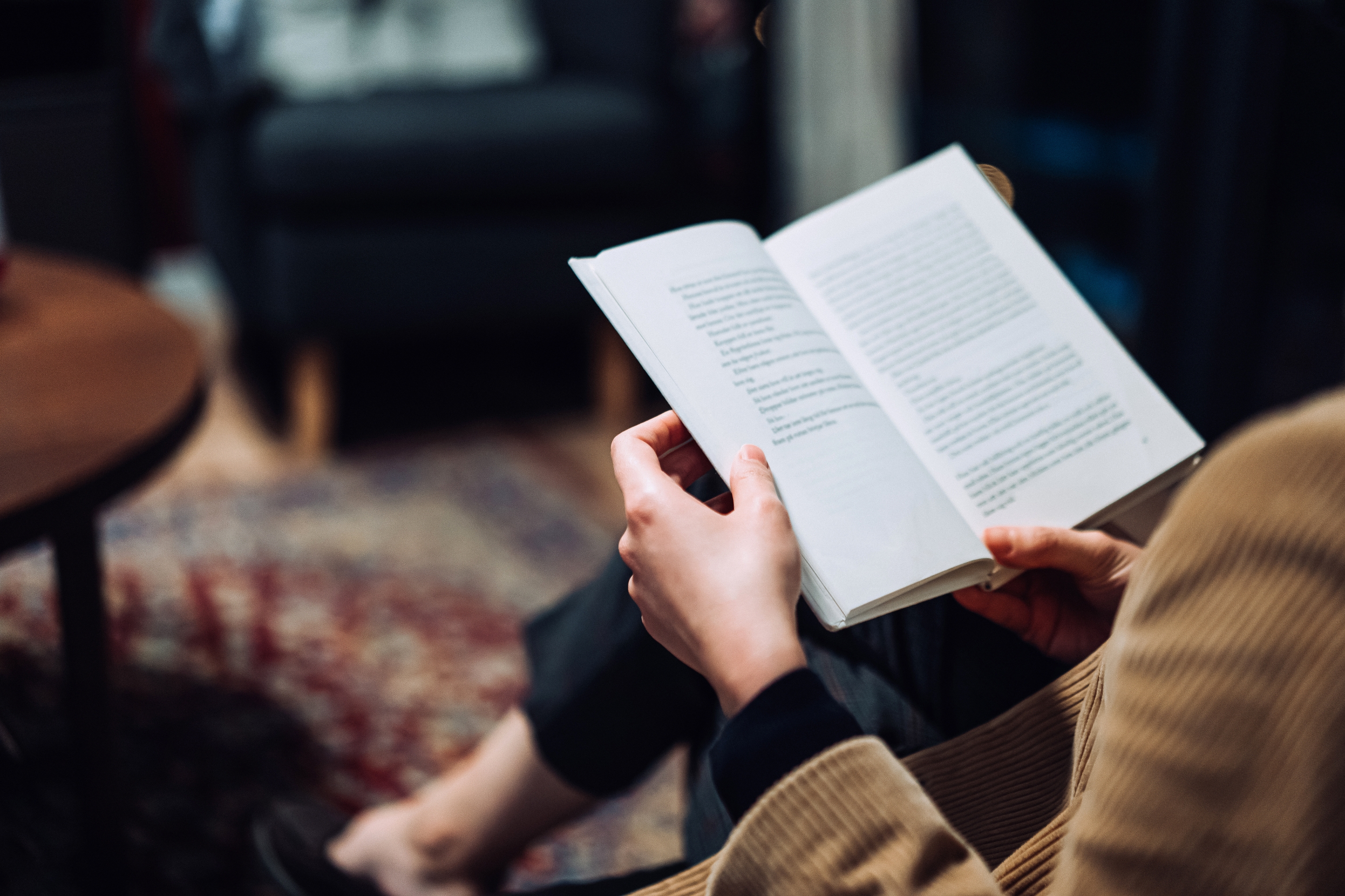 Person reading a book, sitting on a rug with furniture visible in the background, focused on the open pages