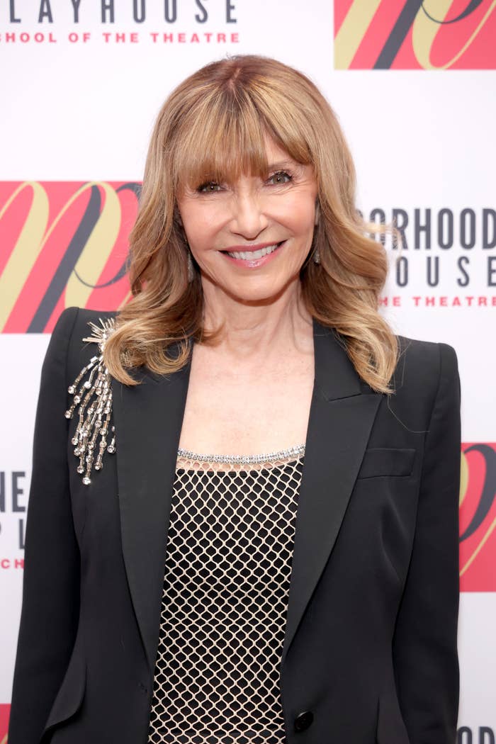 A person smiling on a red carpet, wearing a blazer with a decorative shoulder embellishment over a patterned top