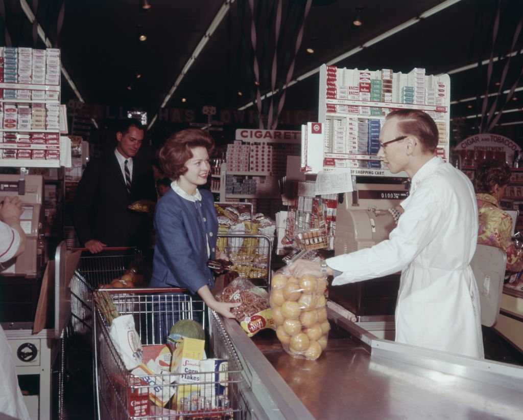 Person at a 1960s grocery store checkout, interacting with a cashier. Shopping cart is filled with various items, including a large bag of oranges