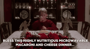 Boy in a festive dining room prays over a plate of macaroni and cheese, surrounded by holiday decorations. Text reads, "Bless this highly nutritious microwaveable macaroni and cheese dinner…"