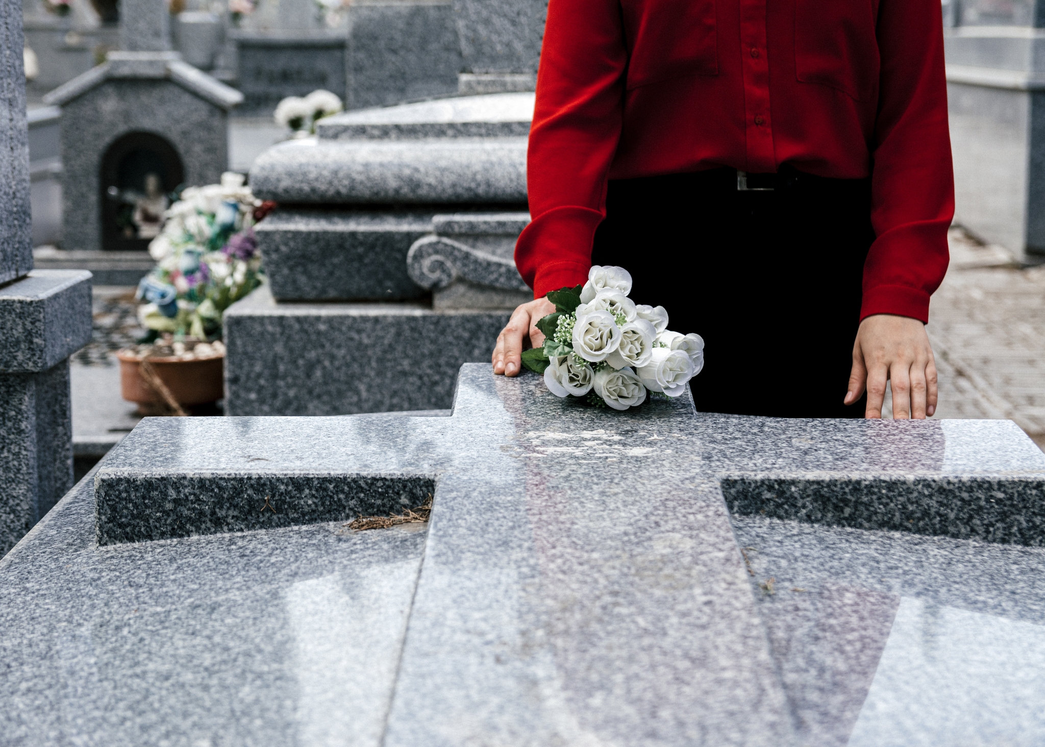 Person in red shirt holding white roses at a grave, surrounded by tombstones and flower arrangements