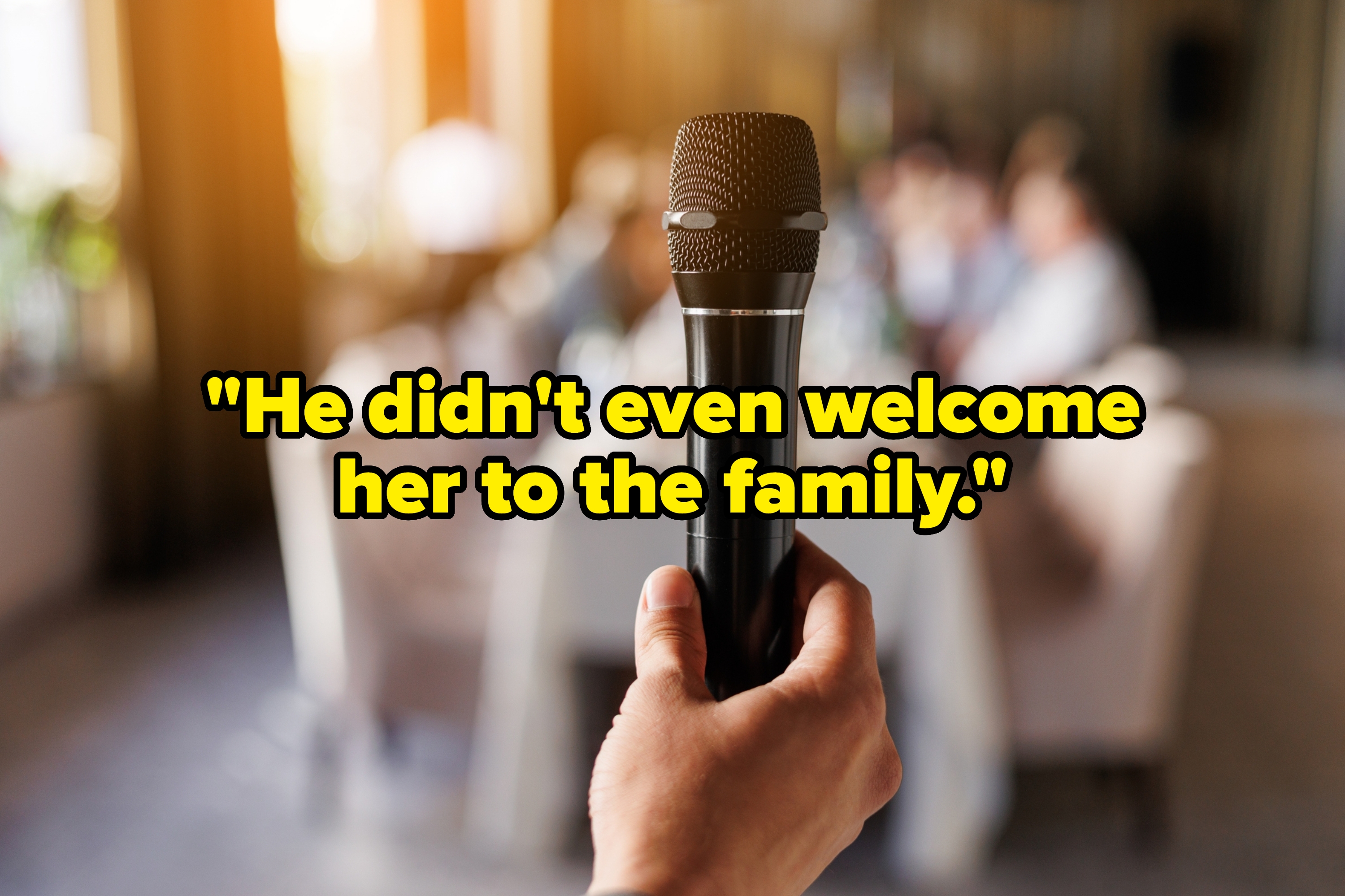 A person holds a microphone, with blurred people in the background seated at a table, suggesting a speech or toast at a wedding event