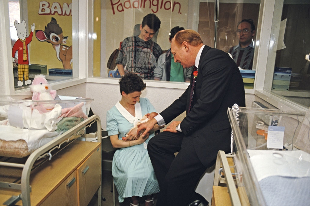 A man wearing a suit talks to a parent holding a newborn in a hospital's maternity ward, with cartoon characters visible on the wall in the background