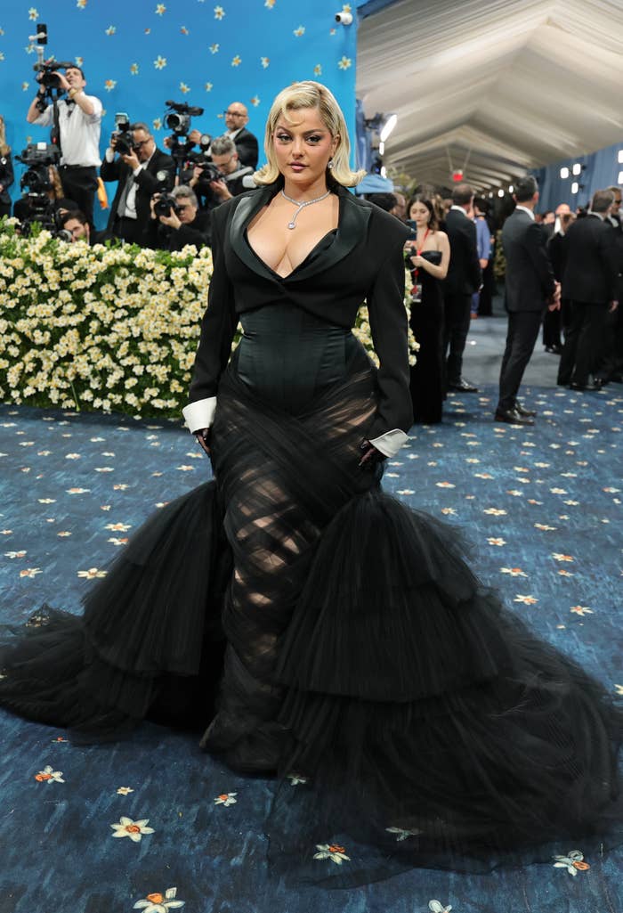 A person in a black, elegant gown with a structured bodice and sheer, flowing skirt poses at a formal event, surrounded by photographers
