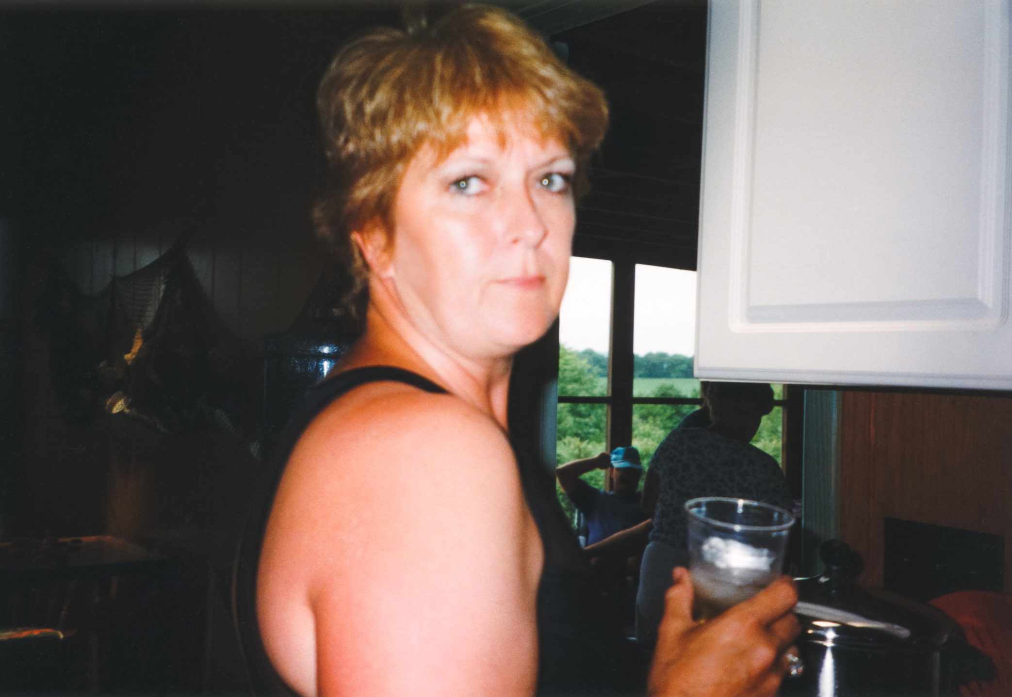 Person in a sleeveless top looks over shoulder, holding a glass with ice, standing in a room with a window view of greenery