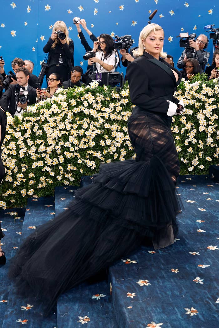 Celebrity in a bold gown with a sheer, layered skirt and a fitted jacket poses on a floral-themed event carpet, surrounded by photographers