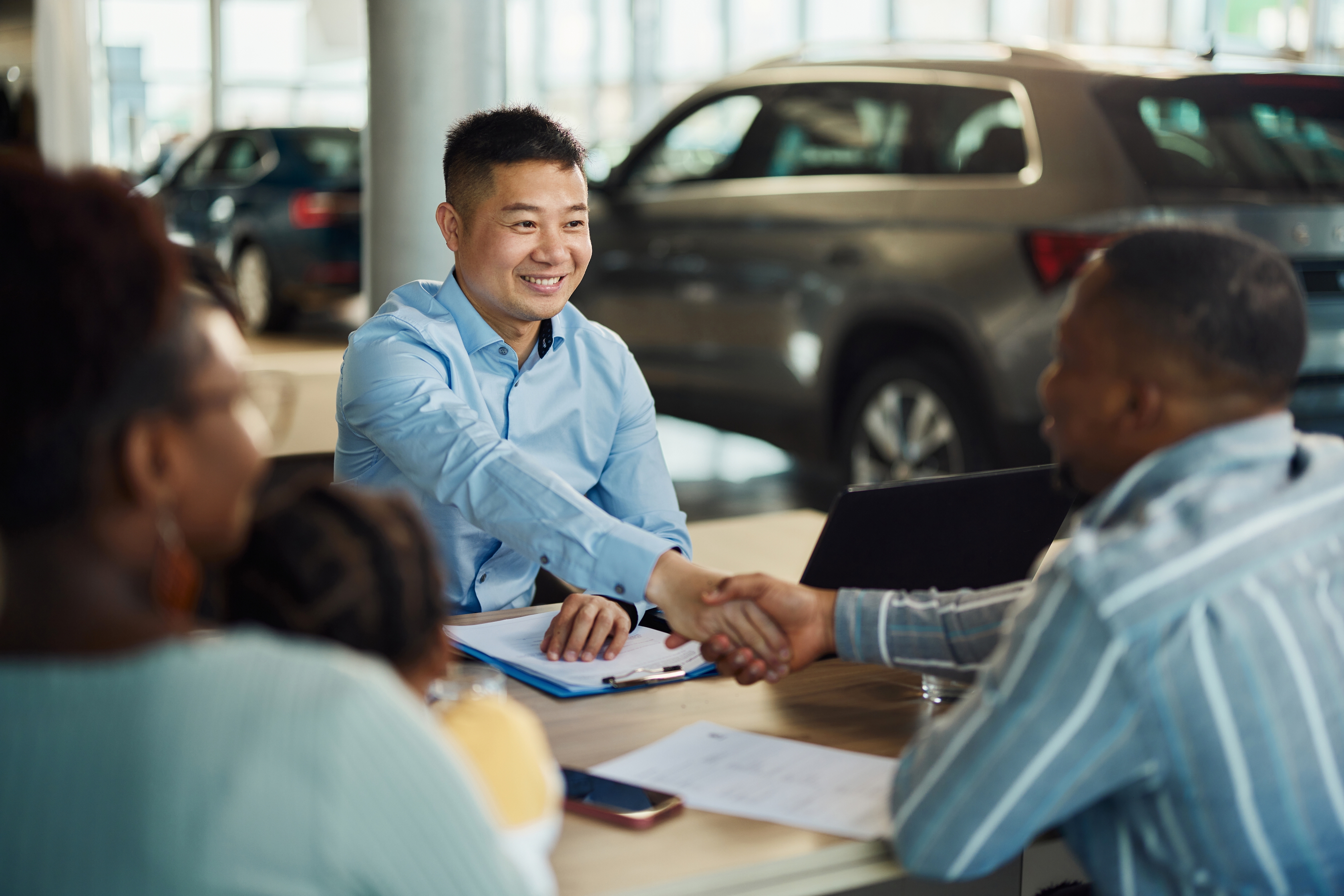 People at a car dealership table, smiling and shaking hands, likely sealing a deal