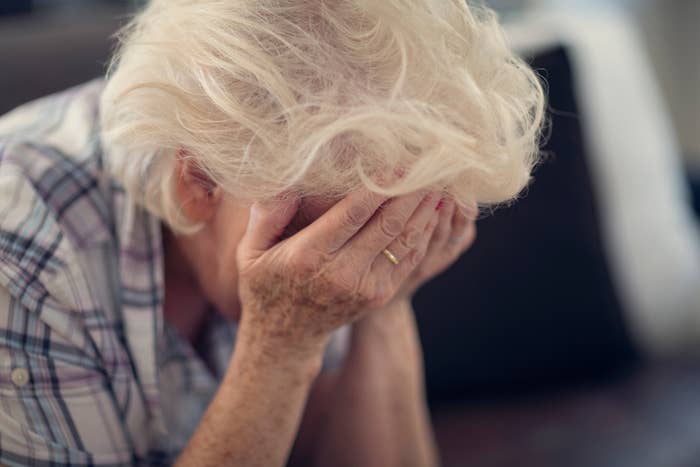 An elderly person with their face in their hands, wearing a plaid shirt and a ring, appears distressed or deep in thought