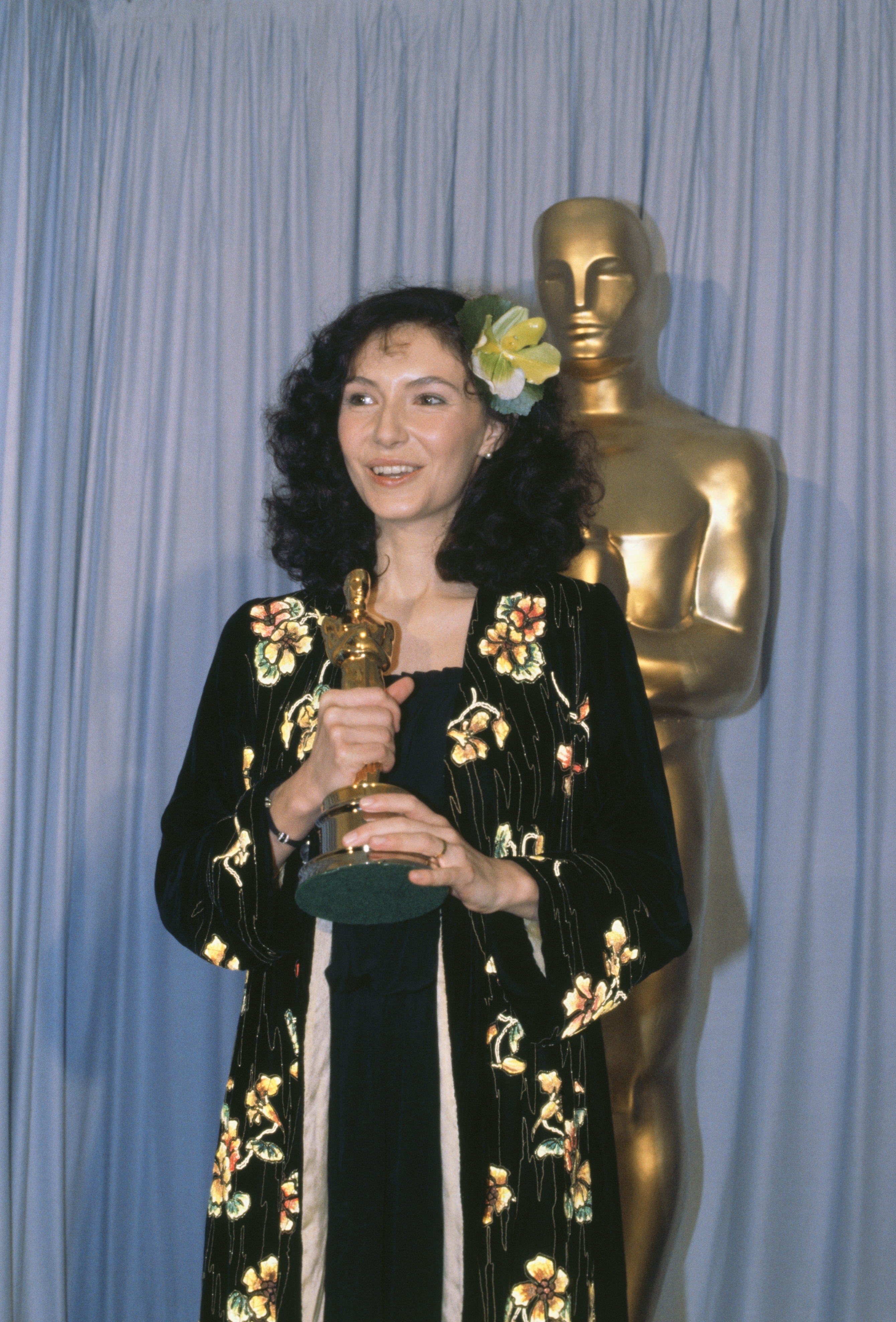 Person holding an award in front of a statue, wearing a floral-patterned velvet outfit with a flower in their hair