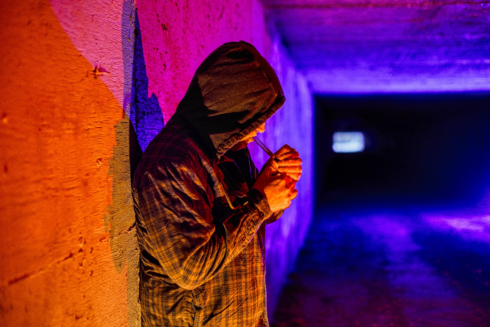 Person in a hooded jacket leans against a wall in a dimly lit tunnel, focusing on lighting an object
