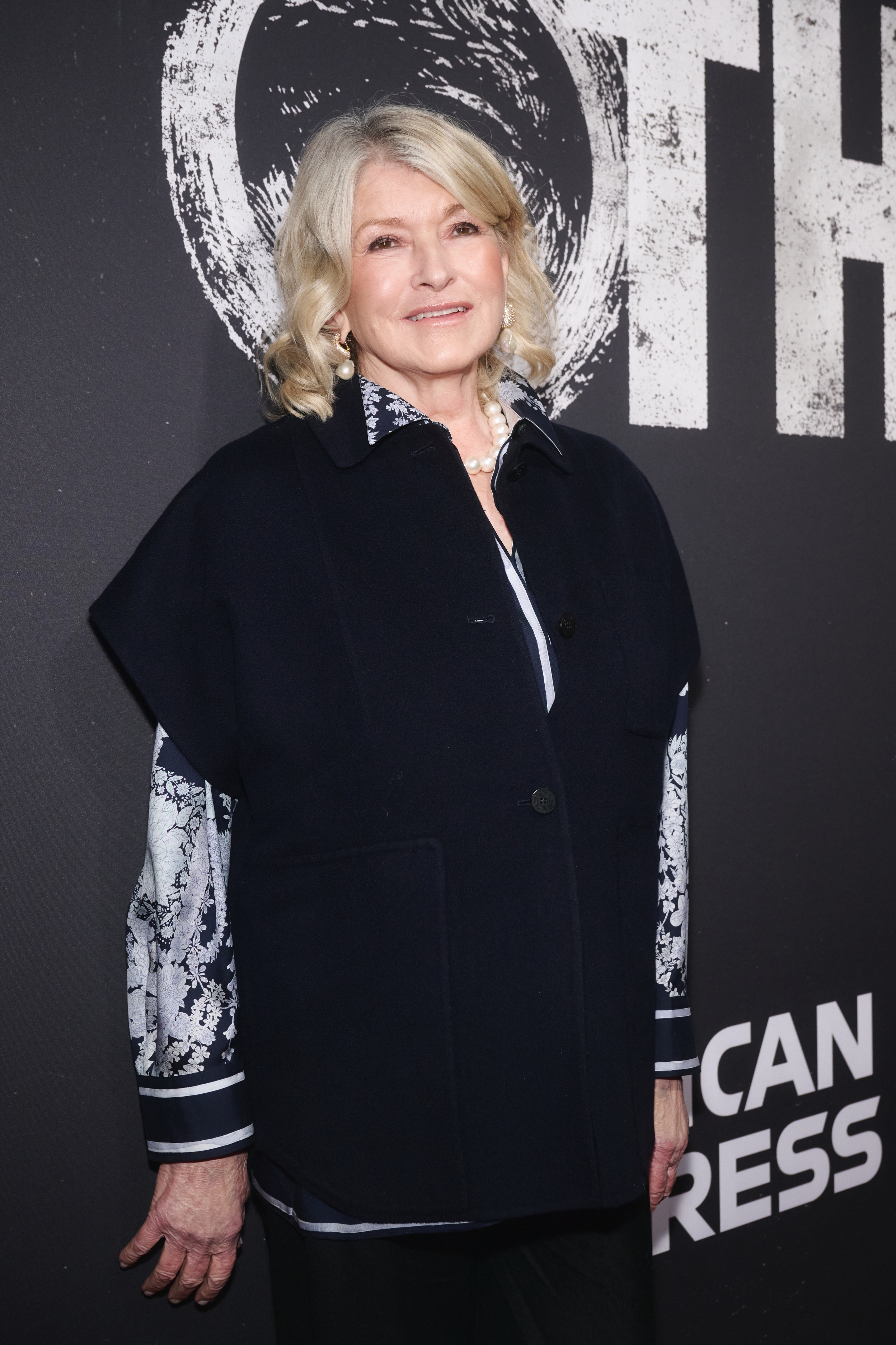 Person in an elegant black coat with a patterned blouse attends a formal event against a backdrop with text