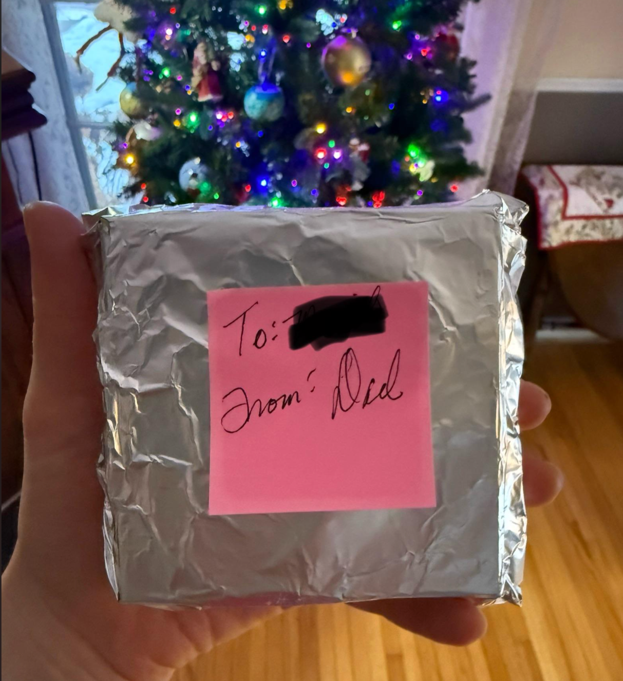 Hand holds a foil-wrapped gift with a pink sticky note, &quot;To: [name redacted], From: Dad.&quot; A decorated Christmas tree is in the background