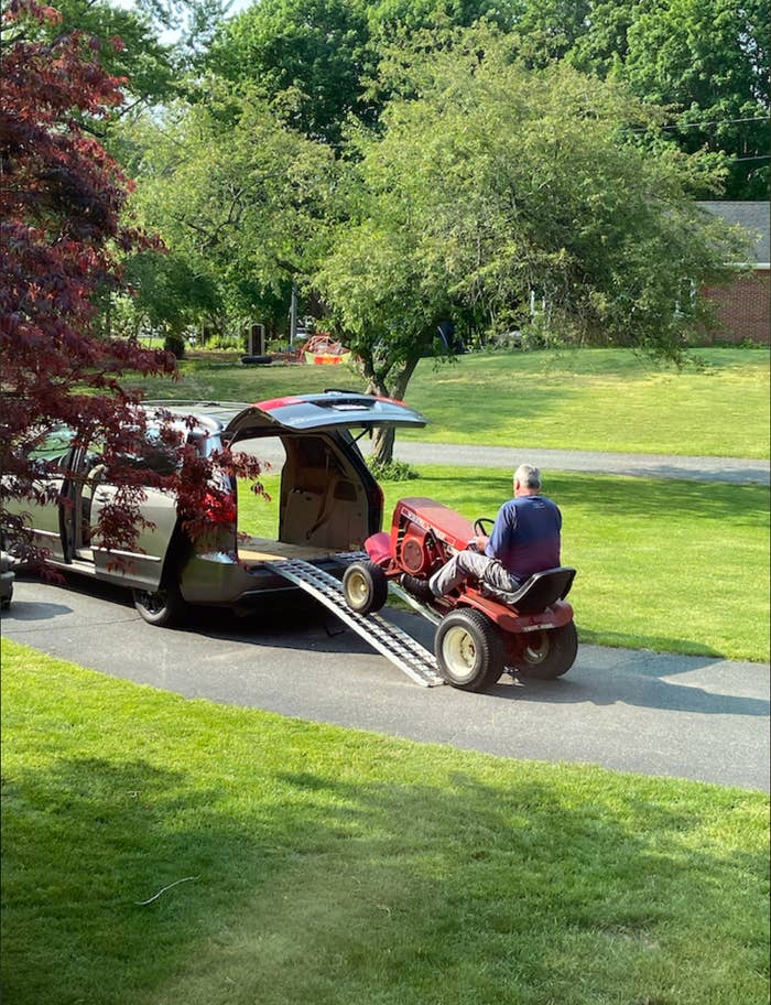 A person drives a ride-on lawnmower up a ramp into a minivan located in a suburban driveway
