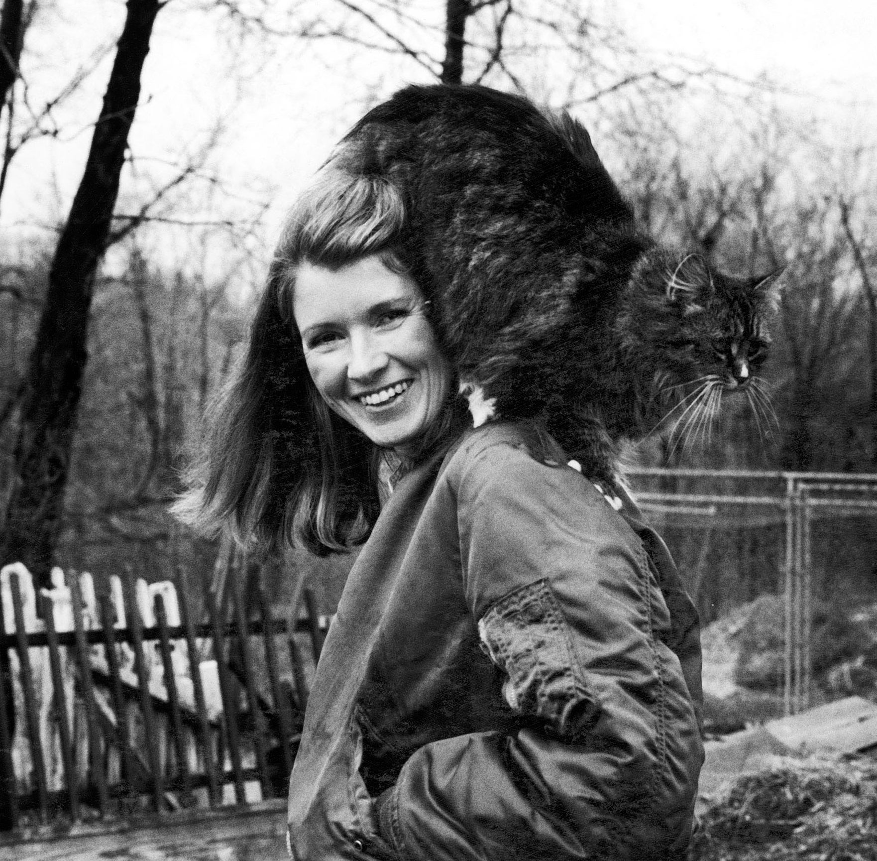 Woman smiling with a large cat perched on her shoulders outdoors, standing in front of bare trees and rustic buildings