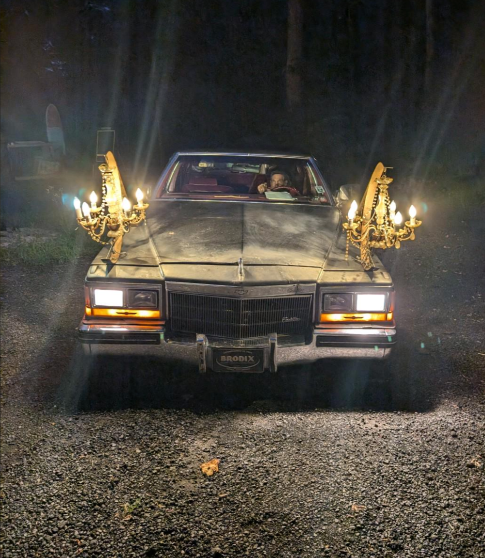 Car with two chandeliers attached to its front, parked on a gravel road at night