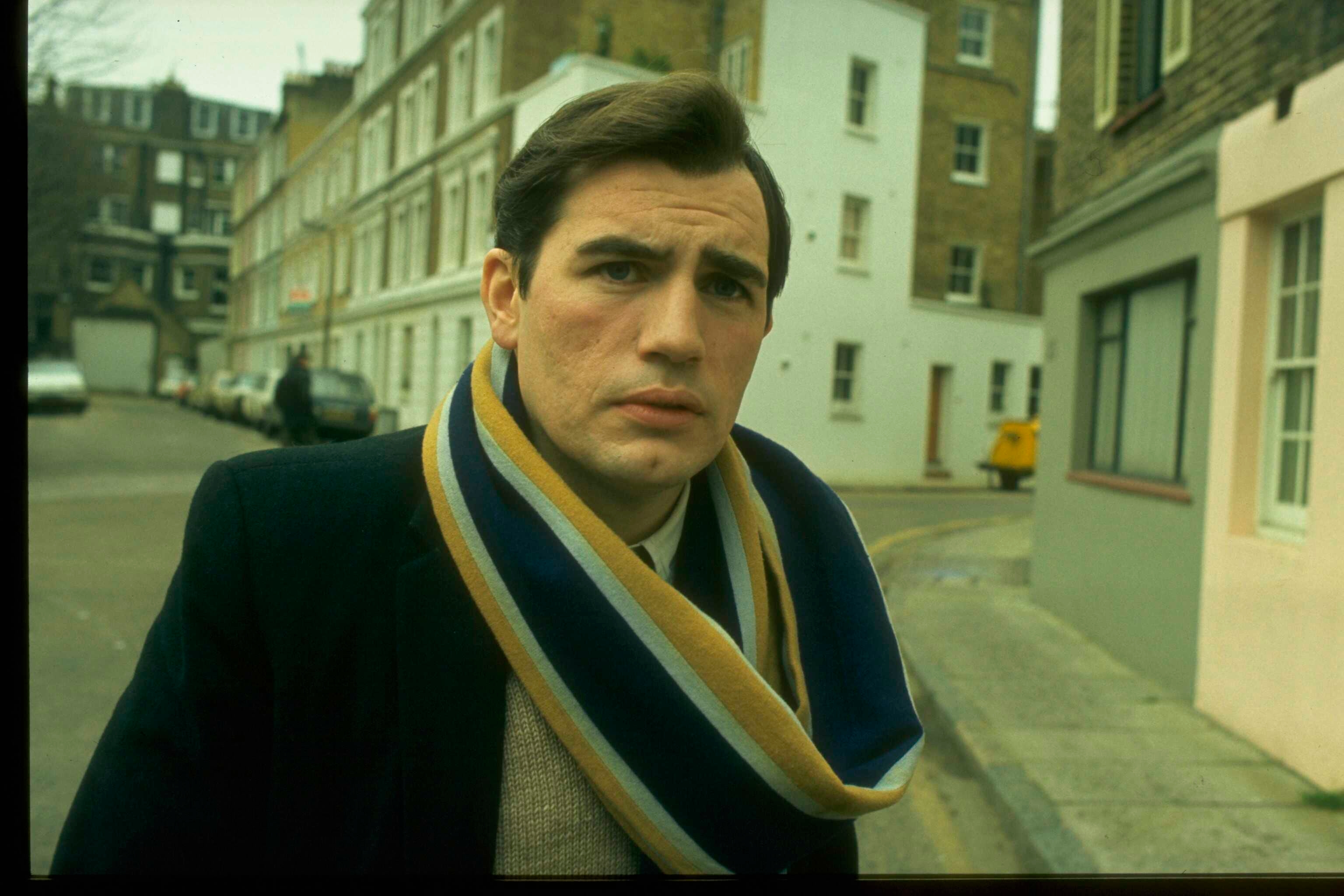 A person standing on a street wearing a dark coat and a striped scarf, looking at the camera with a neutral expression