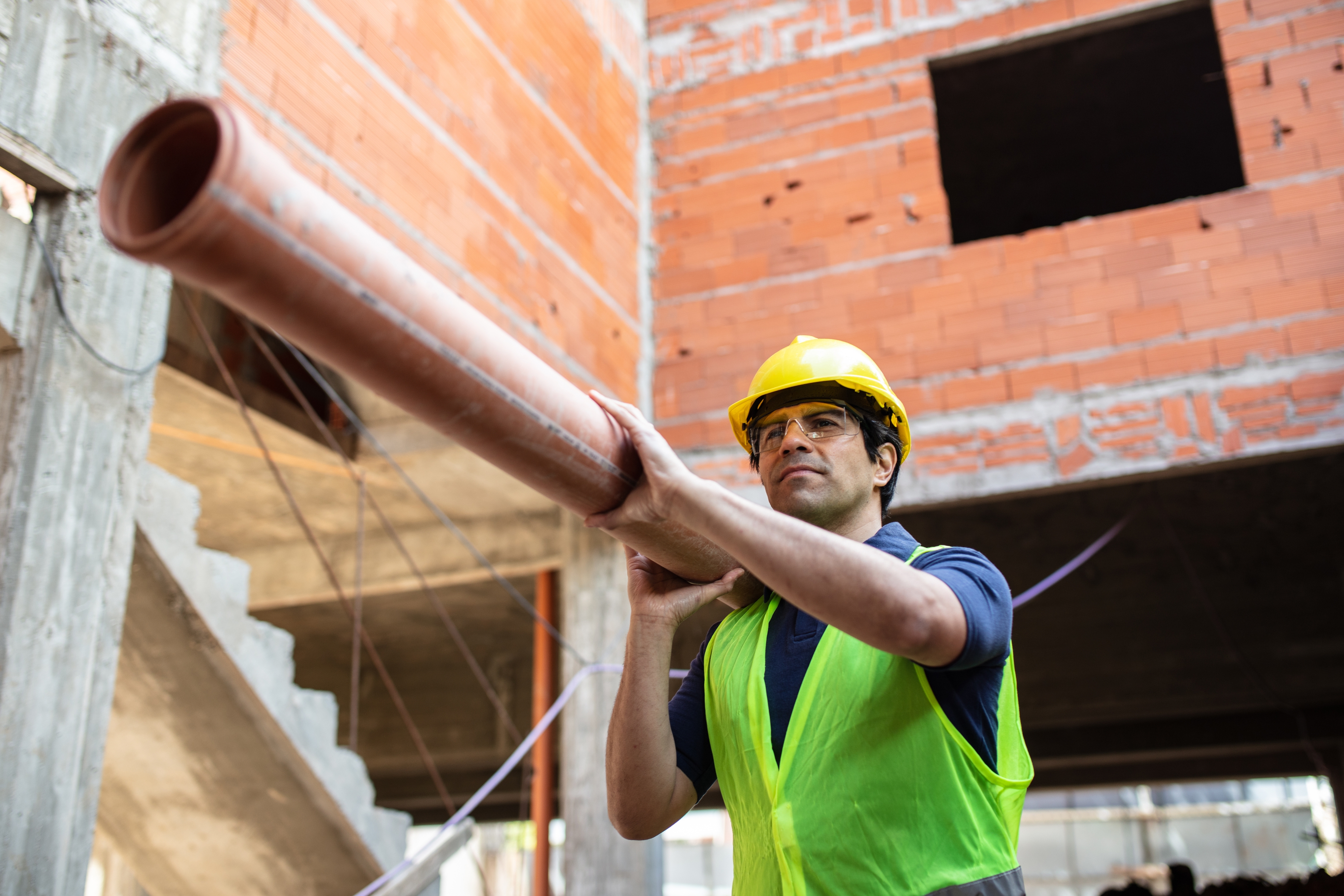 Person in a construction site playfully using a large pipe as a telescope, wearing a hard hat and safety vest