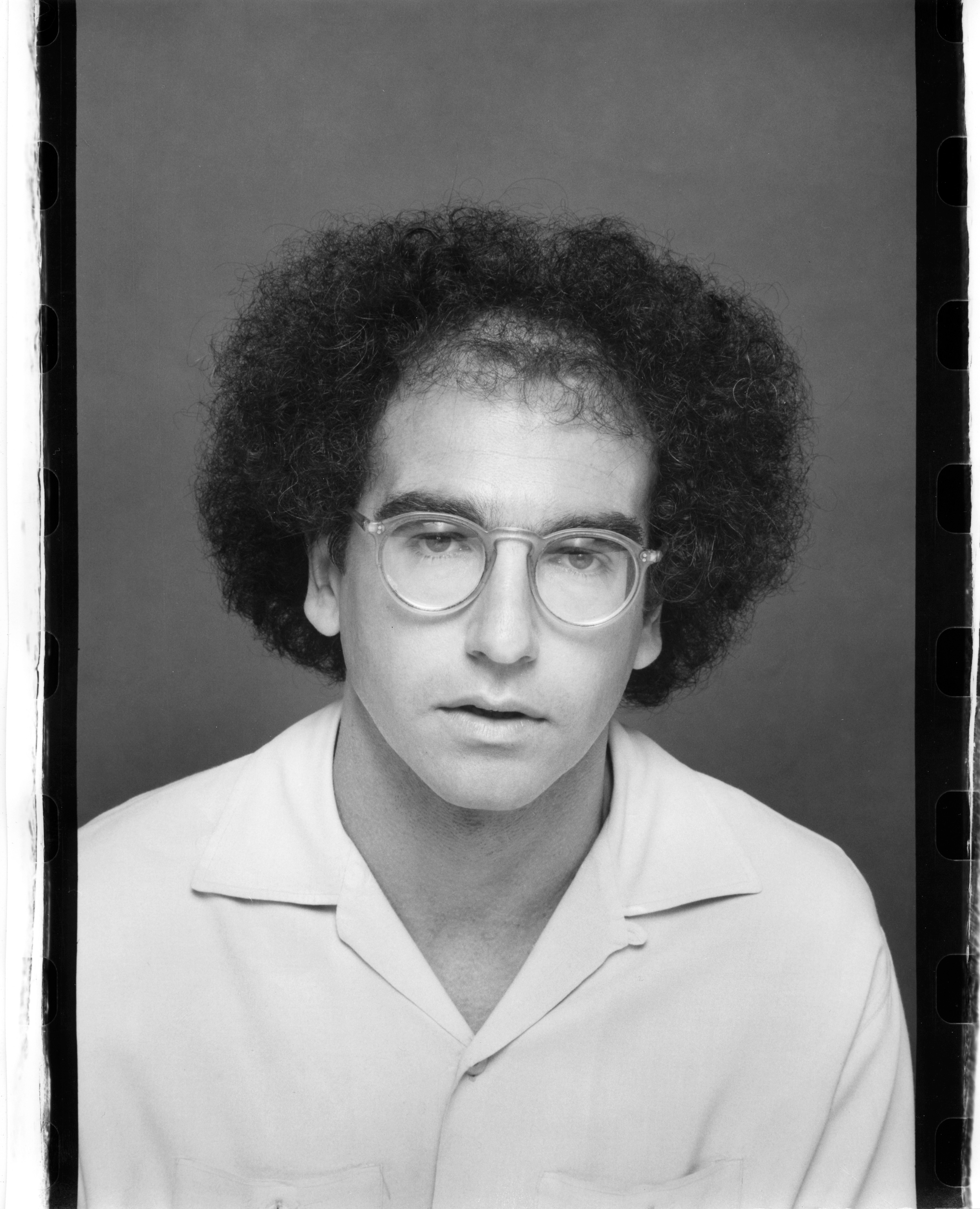 Person with glasses and curly hair, wearing a casual shirt, poses for a formal portrait against a plain backdrop