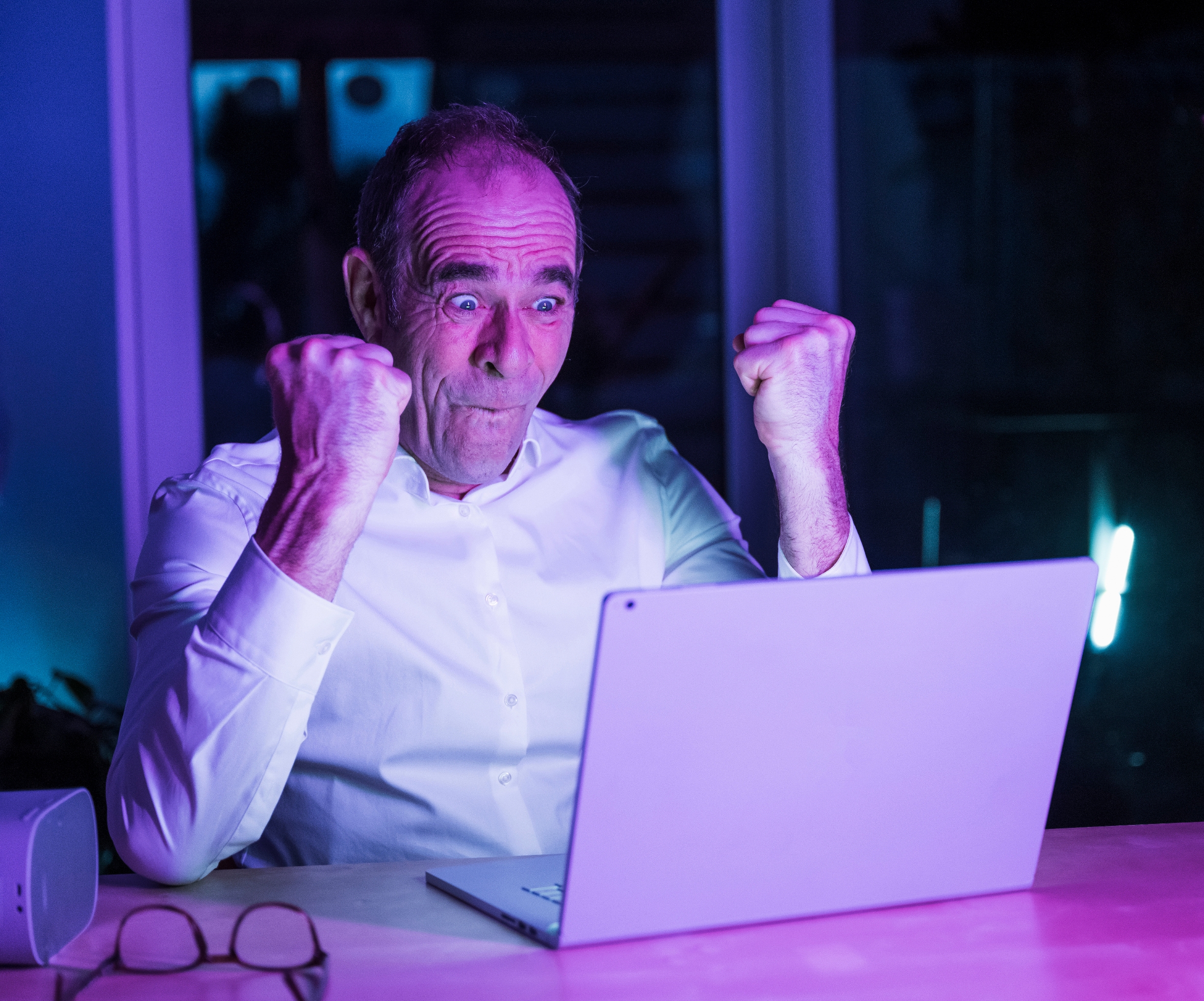Man in white shirt looks excited, raising fists in front of a laptop, possibly in surprise or triumph. Glasses are on the table beside him