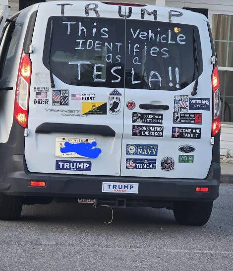Back of a van with bumper stickers and the phrase "This vehicle identifies as a Tesla" written on the window