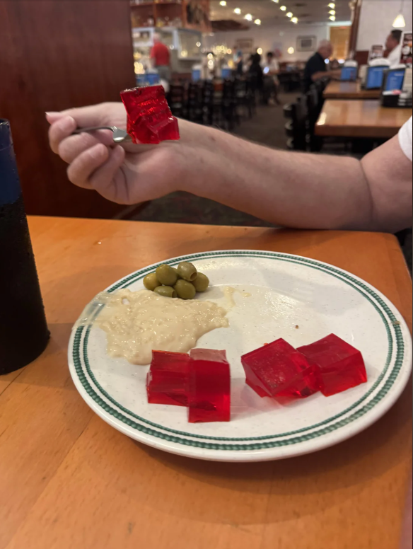 A diner holds a fork with a red gelatin cube over a plate with more gelatin, a beige sauce, and peas. The background shows a restaurant setting