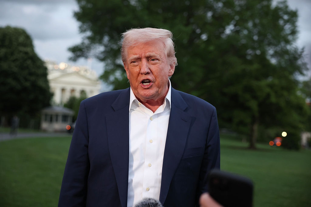 A person in a suit speaks outdoors, surrounded by greenery, with a building visible in the background. A microphone is visible in the foreground