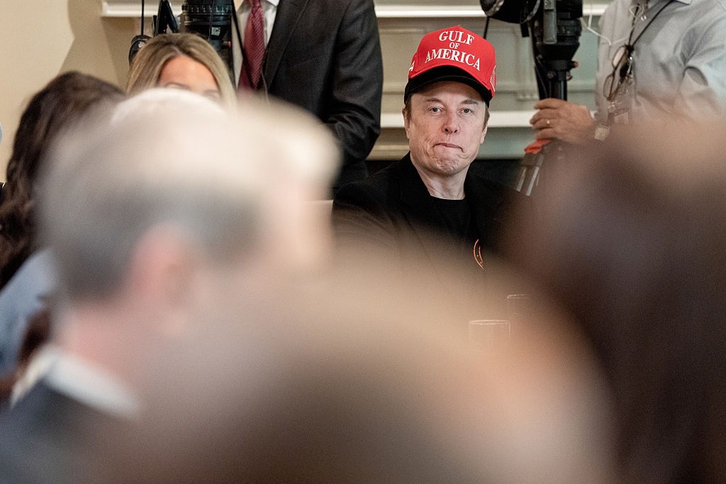 A person in a "Gulf America" cap is sitting among others, appearing focused, at a formal event or meeting with media presence