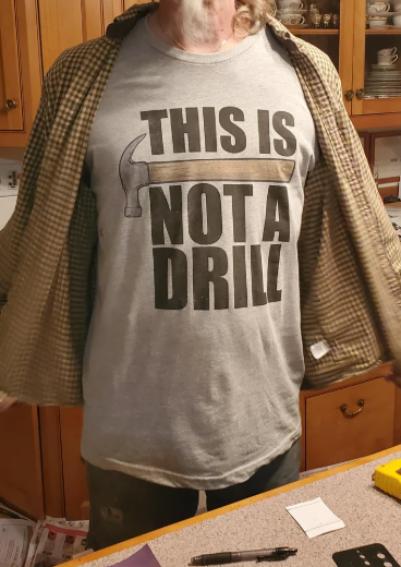 Person wearing a T-shirt with the text "THIS IS NOT A DRILL" alongside an image of a hammer, standing in a kitchen