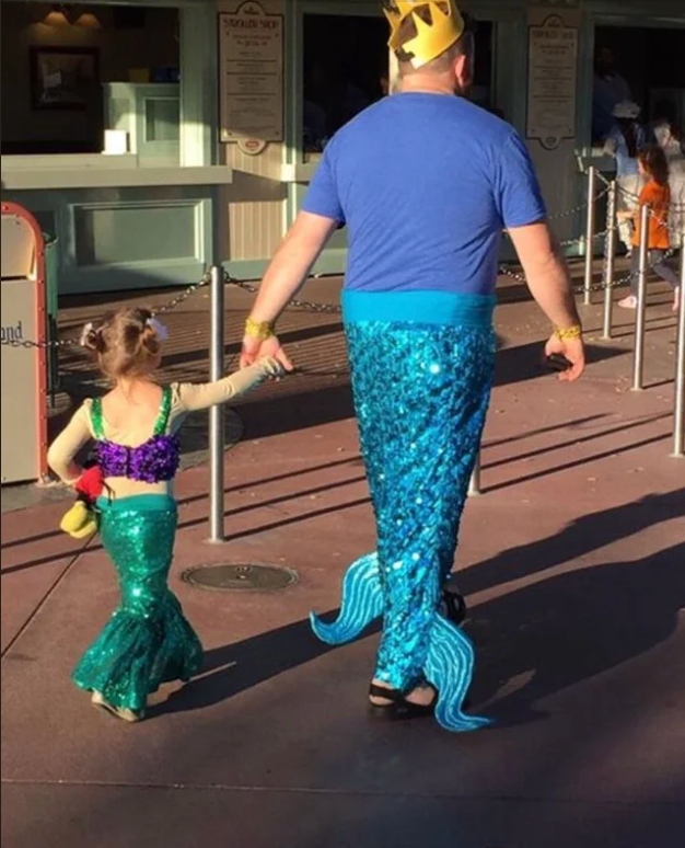 An adult and child walk hand in hand, both dressed as mermaids. The adult wears a crown, while the child holds a plush toy