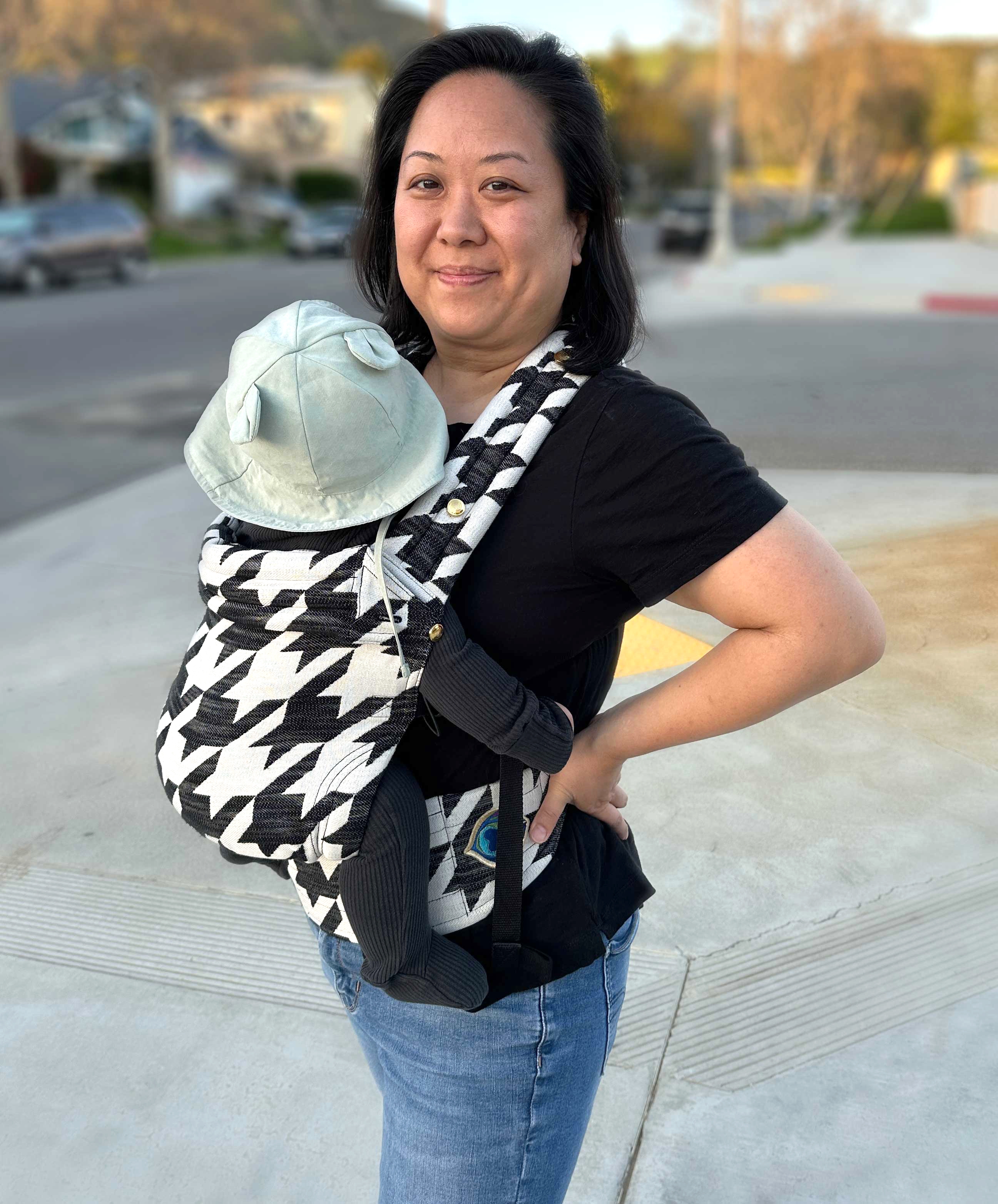 A woman stands outdoors smiling, carrying a baby in a patterned carrier. The baby wears a hat