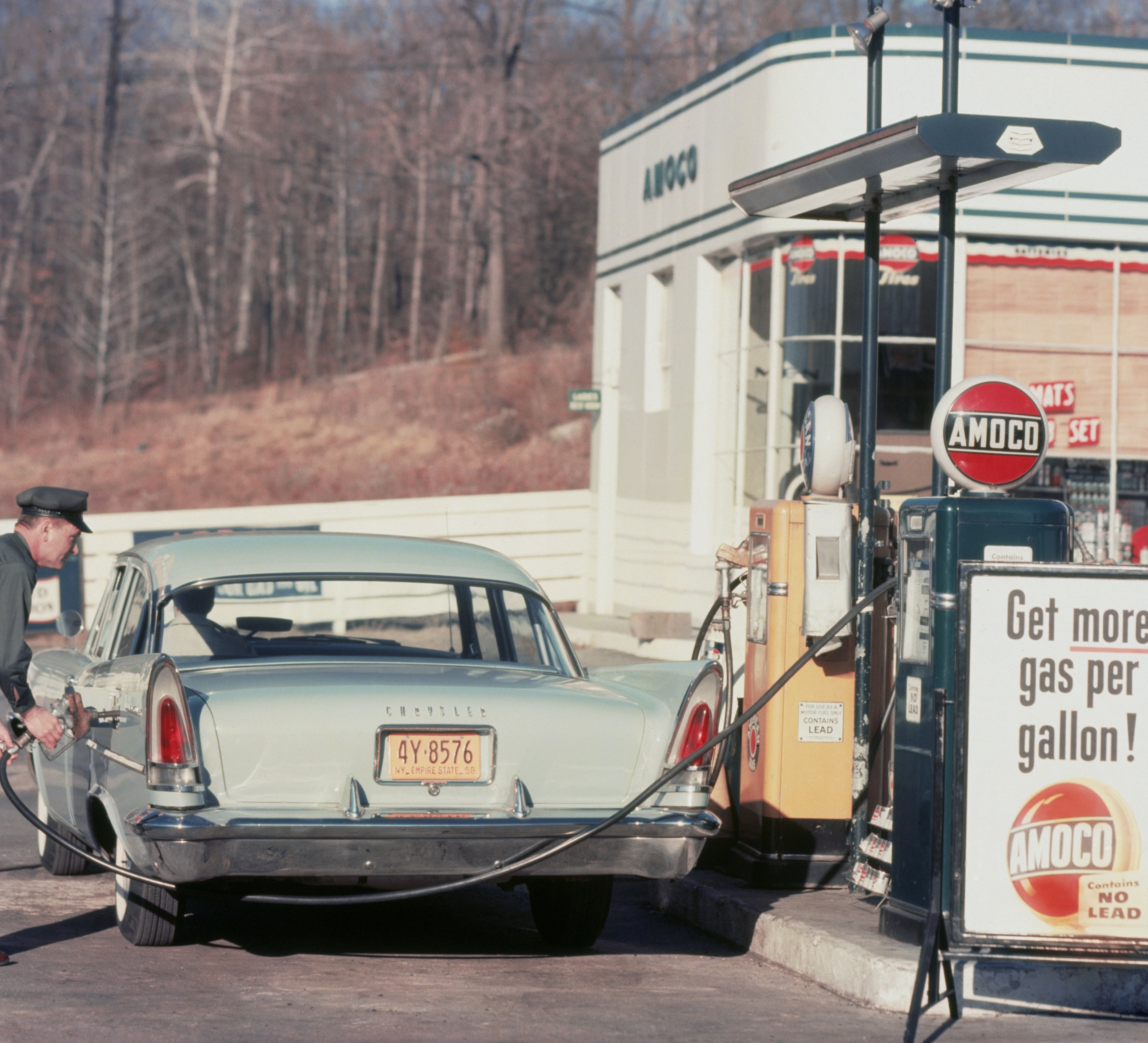 1960s gas station with an attendant fueling a vintage car next to an Amoco pump. Sign reads, "Get more gas per gallon!"