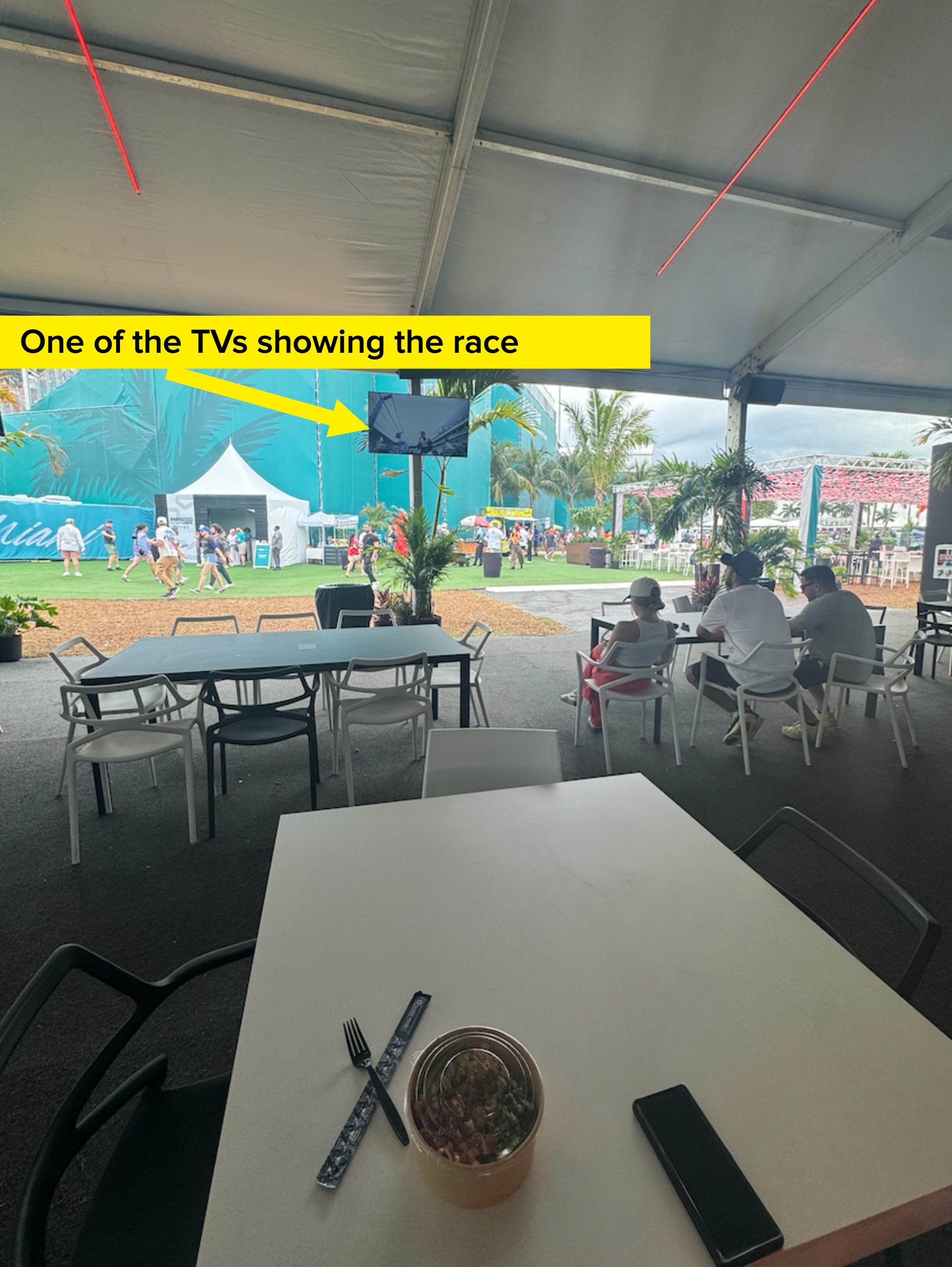 Outdoor dining area under a tent with empty tables, people walking in the background, and a tropical event banner visible