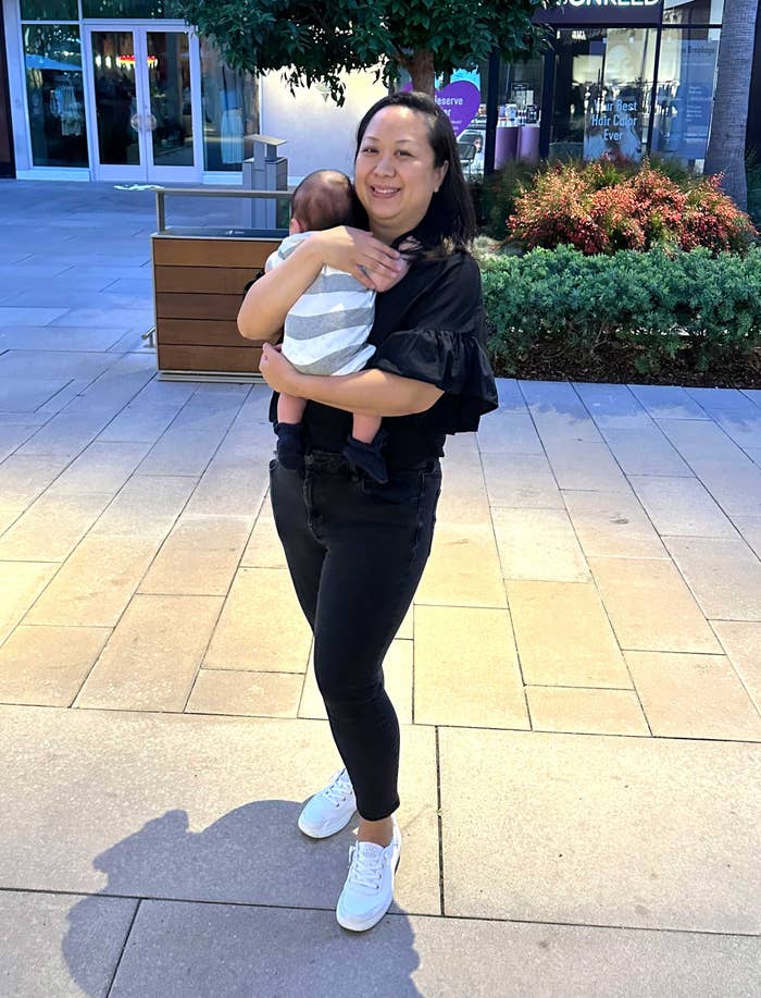 A person in casual attire smiles while holding a baby outside on a paved walkway, with plants and a building in the background
