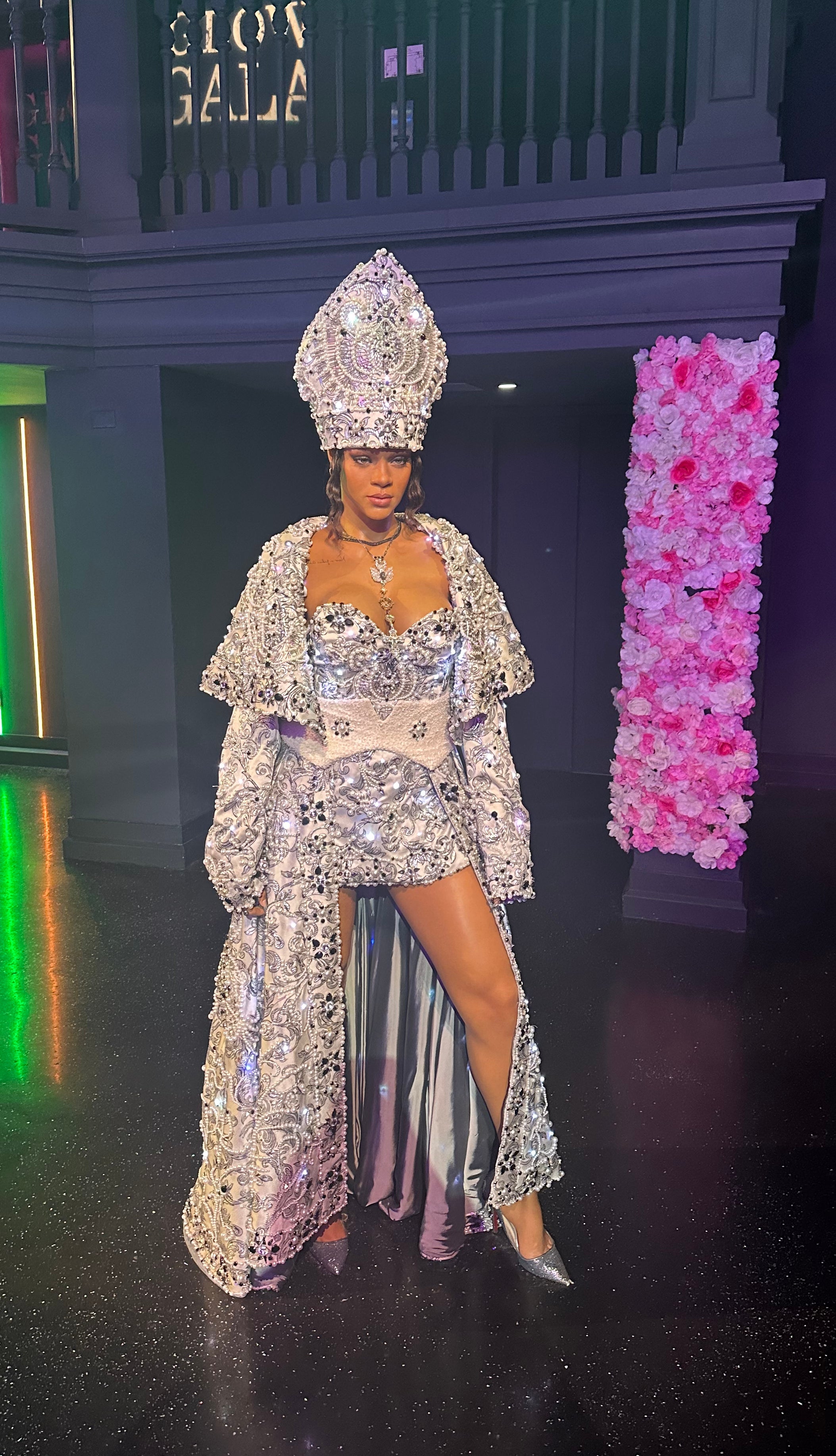 Person in ornate, glittering ensemble with a floor-length coat and headdress, posing confidently at an event with a floral backdrop