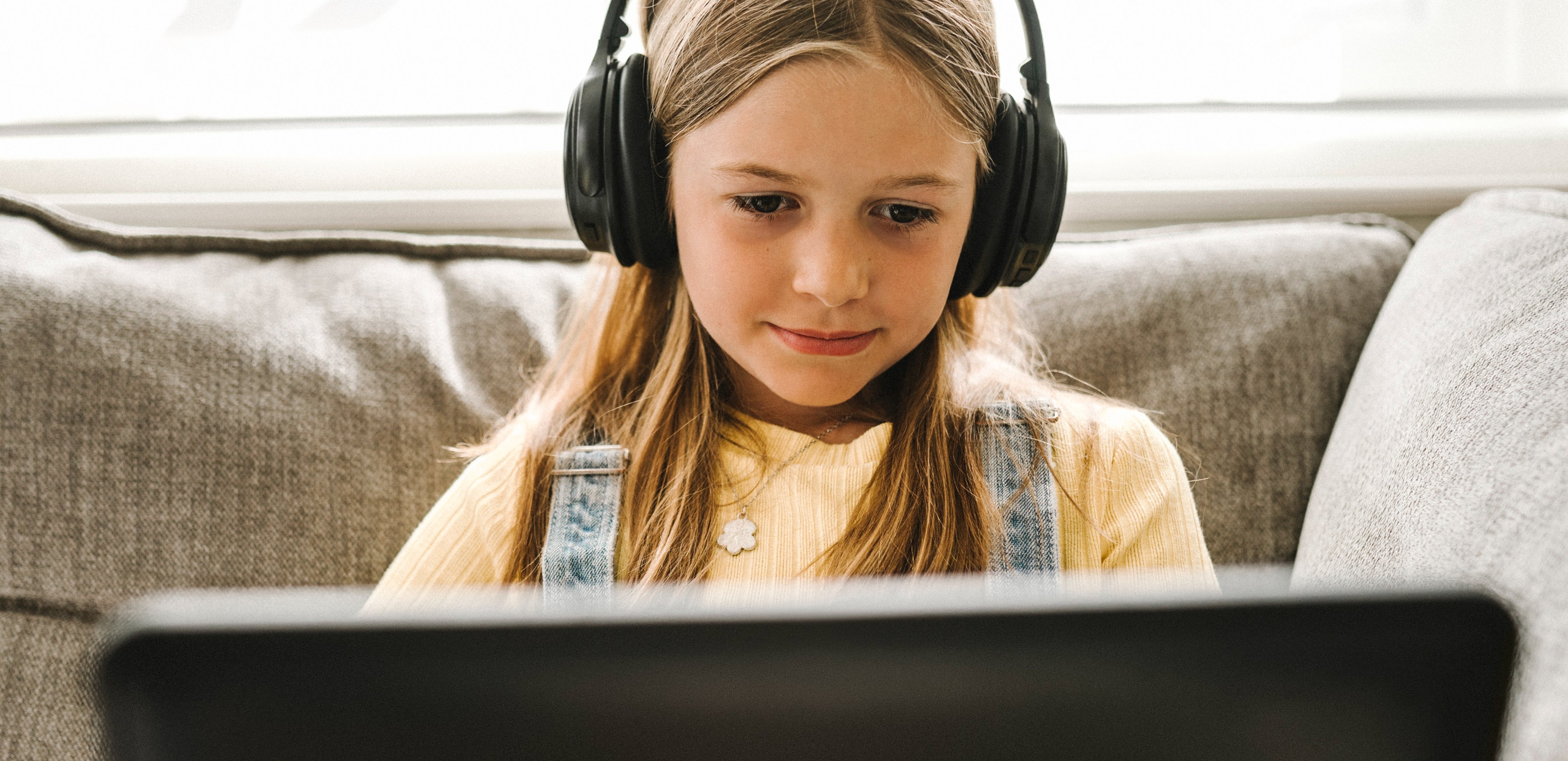 A young girl with headphones sits on a couch, focused on a laptop screen in front of her