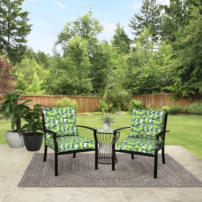 Two patio chairs with leaf-pattern cushions and a tiny array are acceptable connected a mat successful a plot with a woody obstruction and trees successful the background