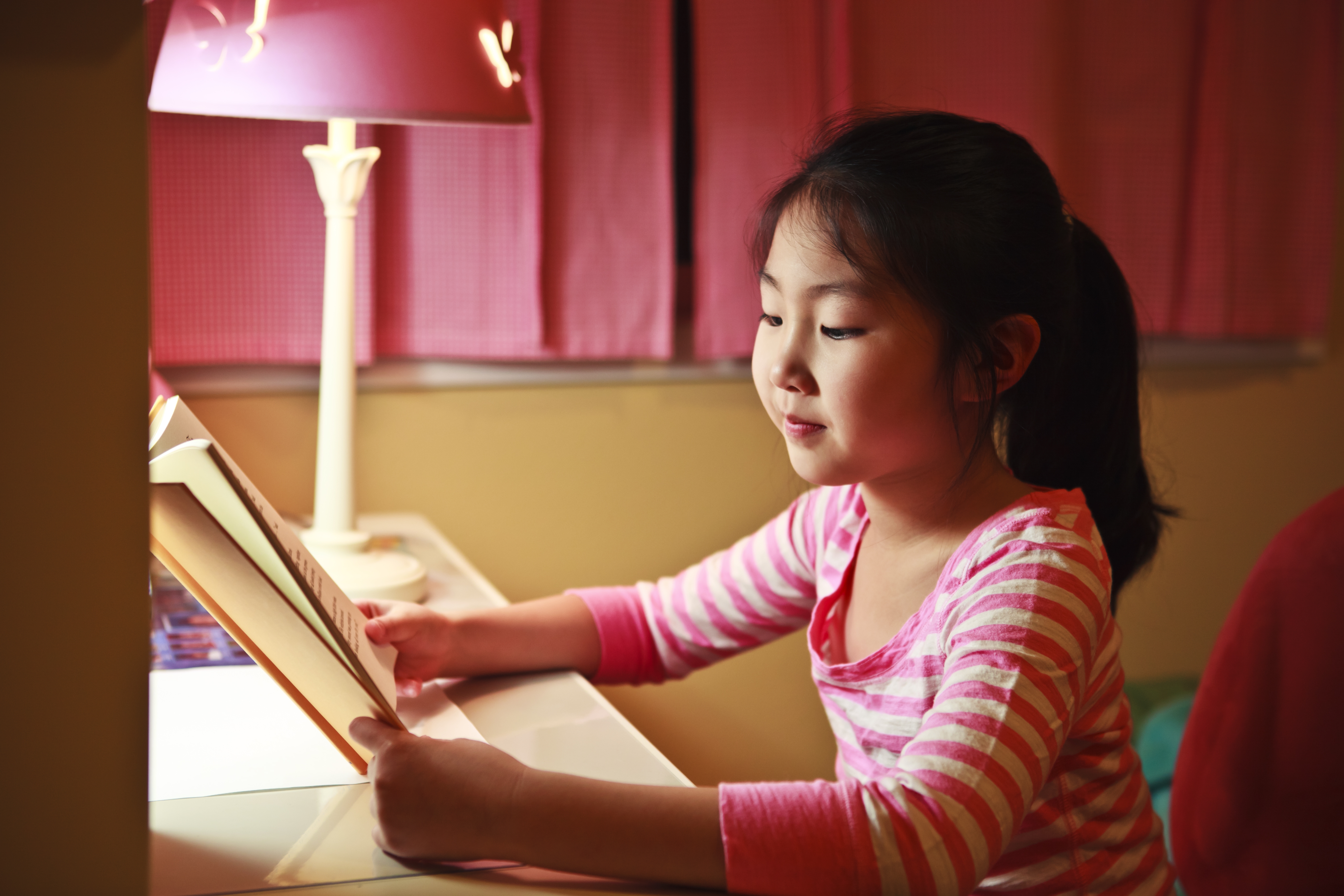Child reading a book at a desk with a lamp nearby, in a cozy, softly lit room