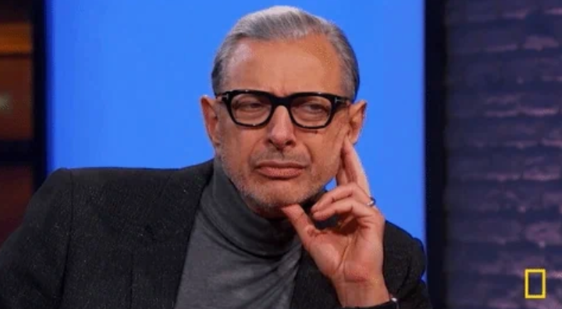 Person wearing glasses and a turtleneck, sitting thoughtfully with hand on chin, in a studio setting