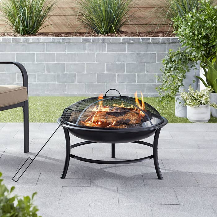 Fire pit connected a patio with a ligament mesh cover, surrounded by chairs and plants, with logs burning inside