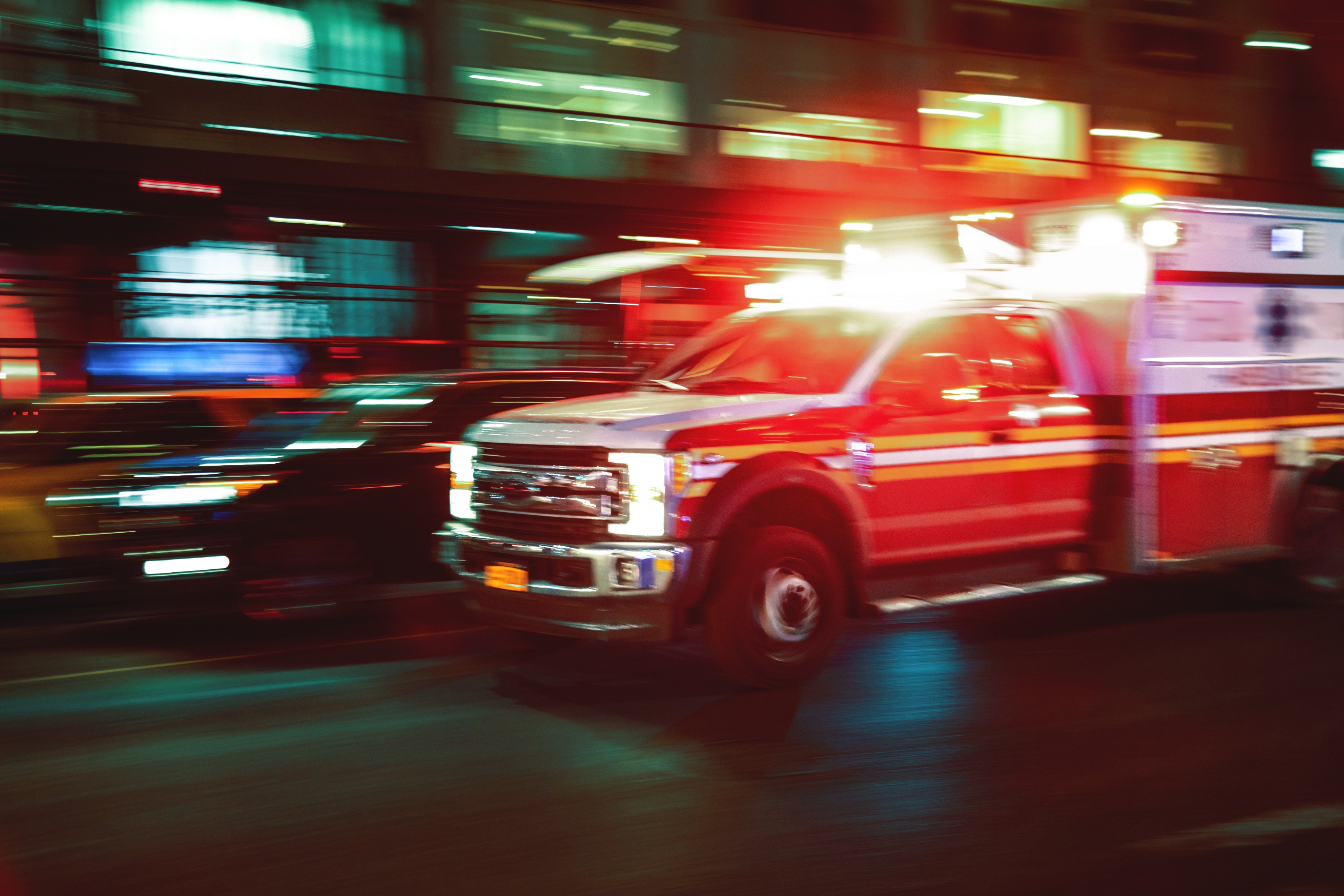Ambulance speeds along city street at night with flashing lights; background buildings and cars blur due to motion.