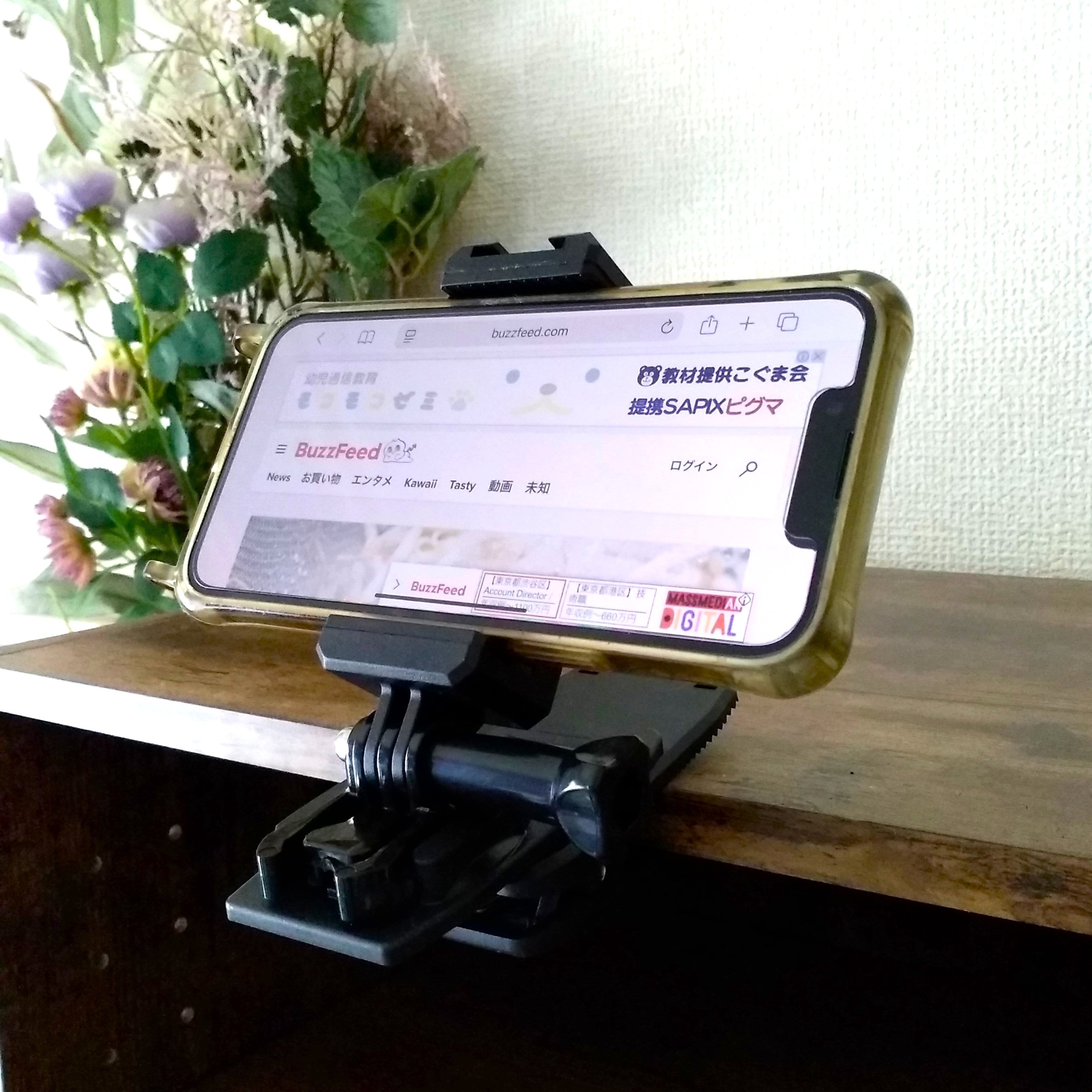 スマートフォンが花の隣の棚にクリップで固定され、画面にBuzzFeedのウェブサイトが表示されています。