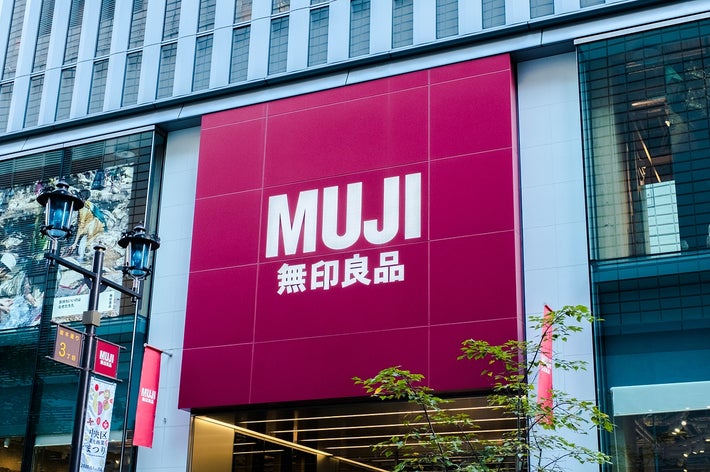 無印良品の店舗外観。大きな看板に「MUJI 無印良品」と表示されています。木や街灯が前景に見えます。