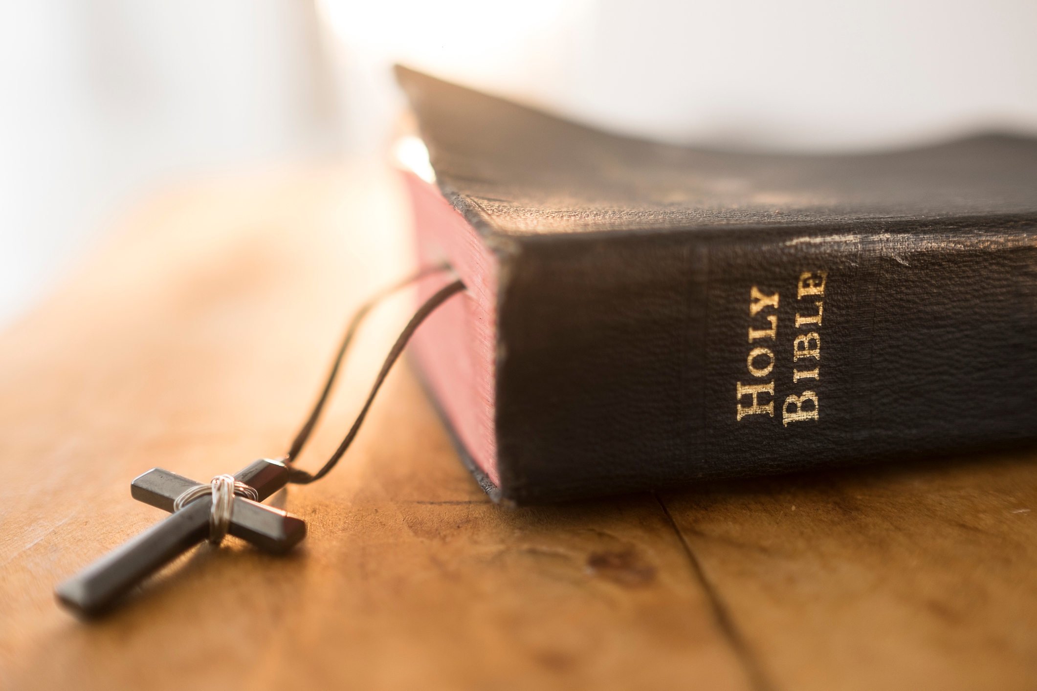 A closed Bible with a bookmark draped over the edge sits on a wooden surface. A small cross is attached to the bookmark