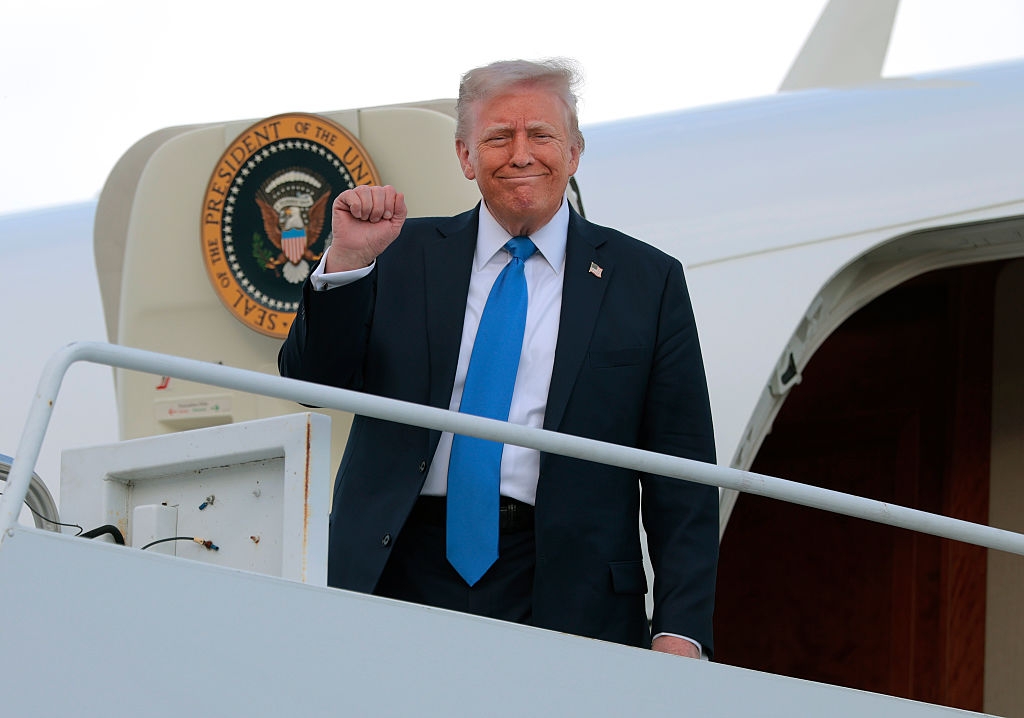 A man in a suit and blue tie stands on stairs near an airplane, raising his fist and smiling. Presidential seal visible on the plane
