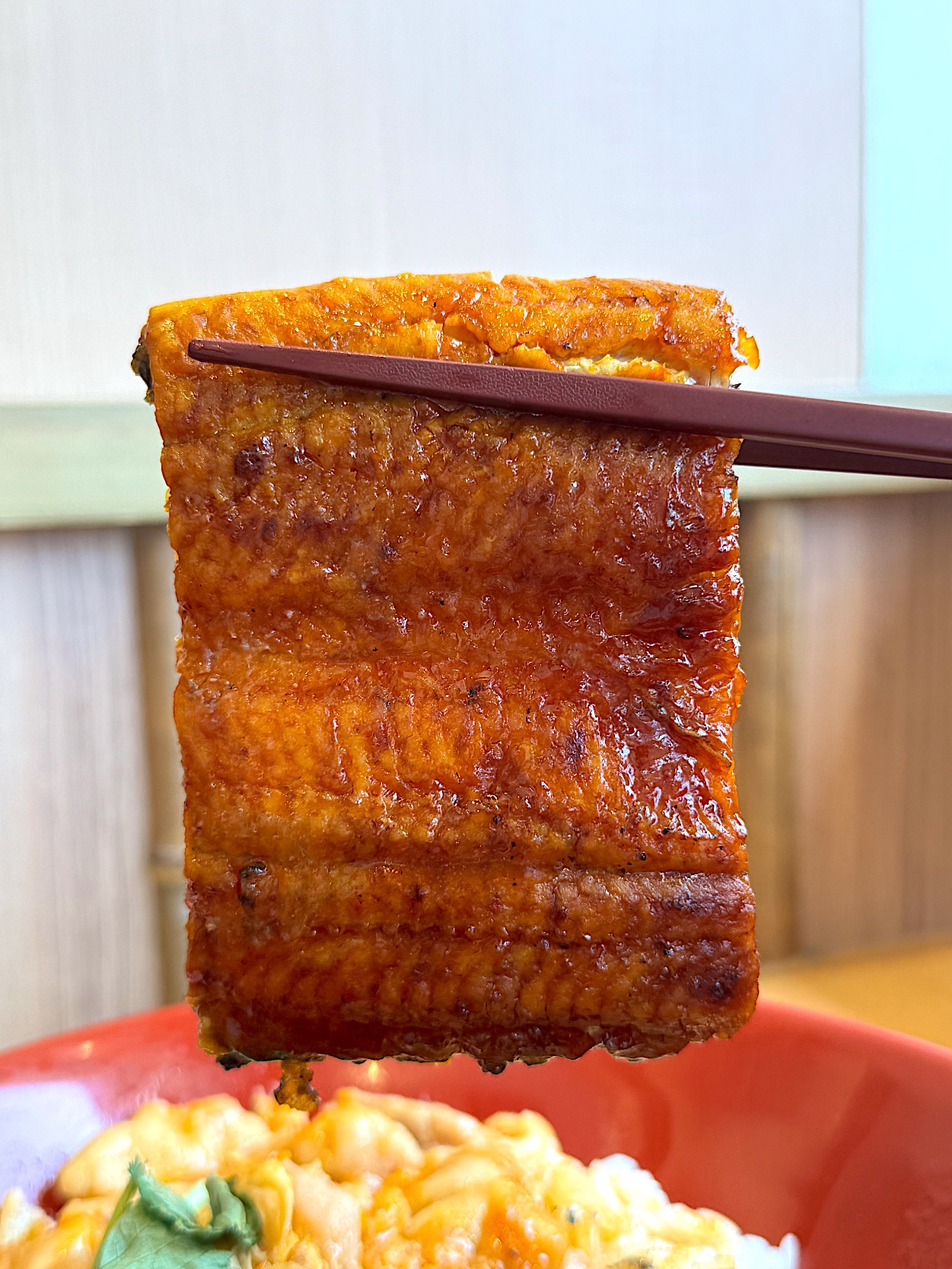 Chopsticks hold up a piece of grilled eel above a bowl of rice and vegetables