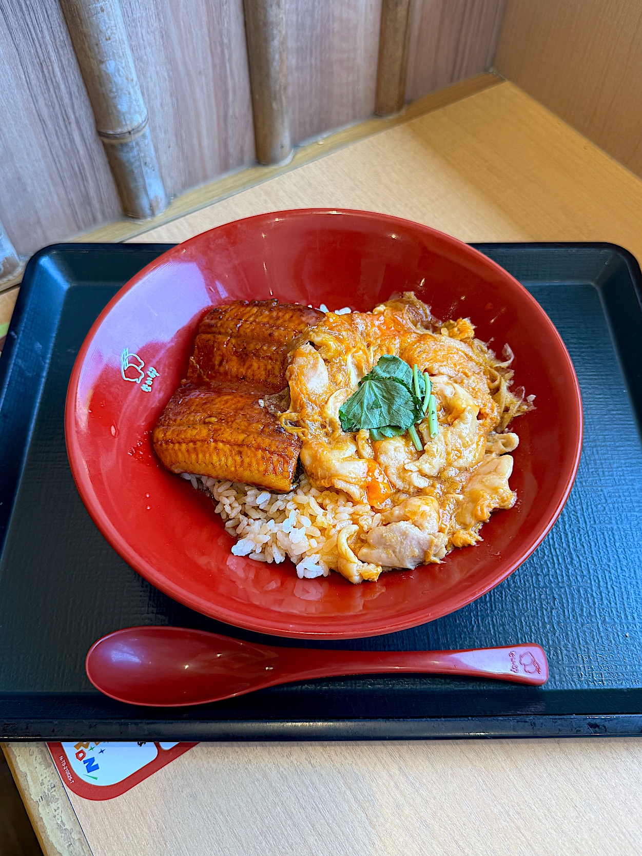 A red bowl of rice topped with cooked eel, egg, and a green garnish is on a tray with a spoon beside it