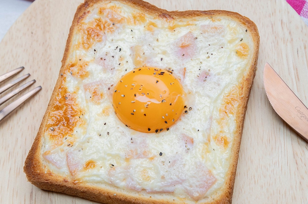 食パンの上にチーズ、ハム、中央に黄身がのった目玉焼きトーストが置かれています。横にはフォークとナイフがあります。