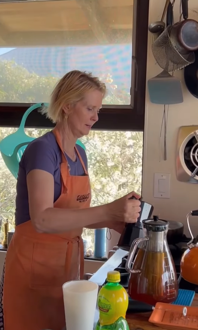 Cynthia Nixon in a kitchen wearing an apron, using a coffee machine