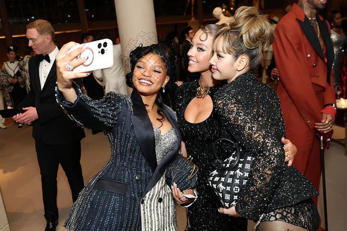 Three people pose together for a selfie at a glamorous event, dressed in elegant evening wear with ornate details